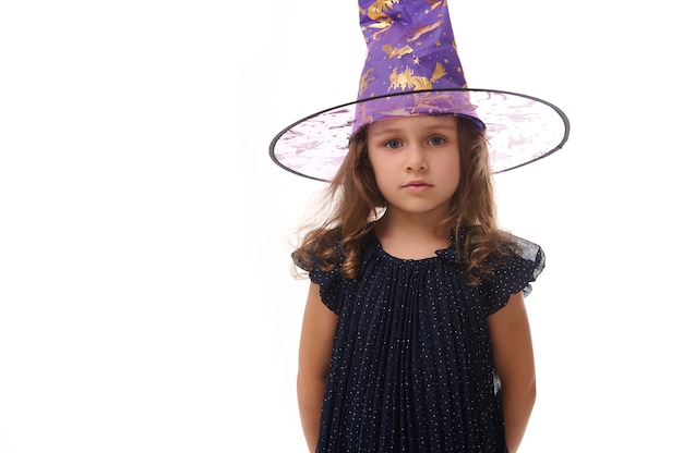 Portrait of beautiful confident little girl wearing a wizard hat and dressed in stylish carnival dress, looking at camera posing with crossed arms against white background, copy space. Halloween