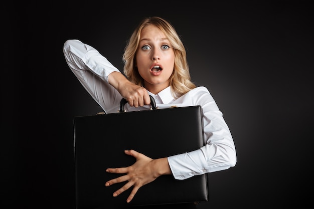 Portrait of a beautiful confused blonde haired business woman dressed in formal clothes standing isolated over black wall, holding briefcase