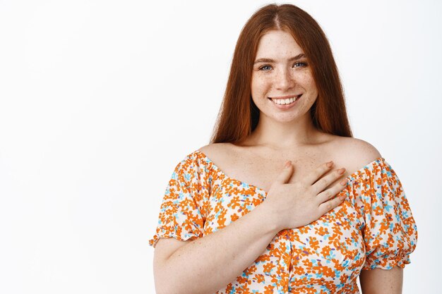 Ritratto di bella ragazza rossa formosa che tiene la mano sul suo cuore che sorride e che guarda felice alla macchina fotografica che sta in vestito sopra priorità bassa bianca