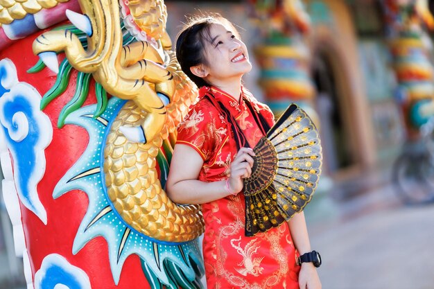 Portrait beautiful cute smiles Asian young woman wearing red traditional Chinese cheongsam decoration and holding a Chinese Fanning for Chinese New Year Festival at Chinese shrine in Thailand