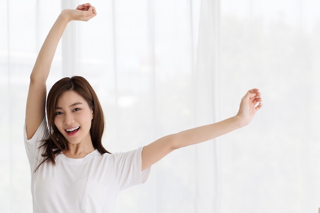 Портрет красивой милой молодой азиатской женщины, улыбающейся уверенно и изящно. Она поднимает руку, чтобы коснуться подбородка в дружеской позе на белых занавесках.