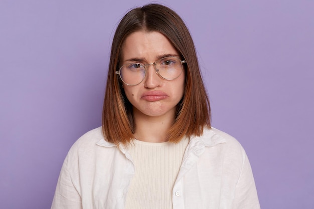 Portrait of beautiful dark haired young Caucasian woman wearing white shirt looking at camera with pout lips being sad and upset expressing sorrow posing isolated over purple background