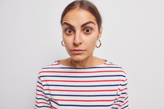 Portrait of beautiful dark haired young woman looks with wonder at camera hears shocking news finds out something interesting raises eyebrows from surprisement poses against white background.