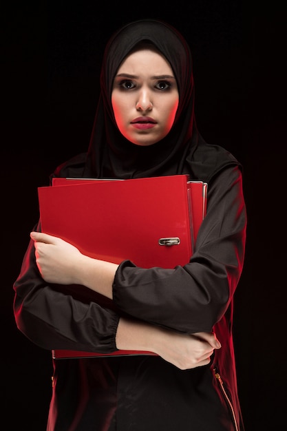 Portrait of beautiful desperate scared frightened young muslim woman wearing black hijab holding folders as keeping secret concept  black  