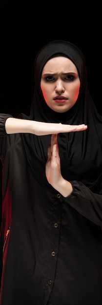 Portrait of beautiful desperate scared frightened young muslim woman wearing black hijab showing stop sign on black