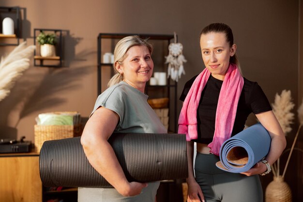 Portrait of beautiful elderly mother and young cheerful daughter wearing sportswear holding mat