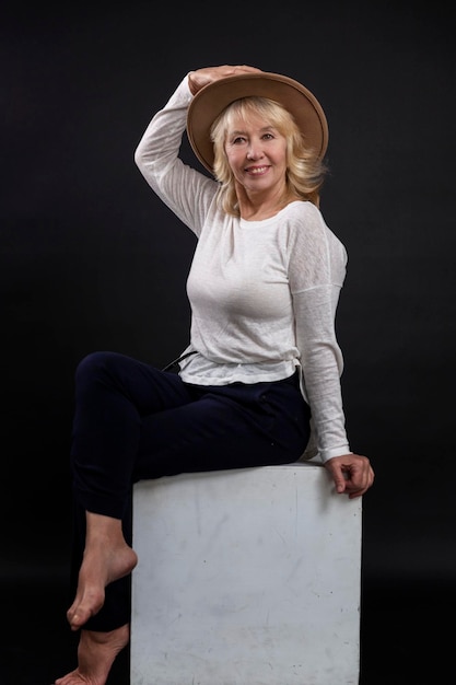 Portrait of a beautiful elderly woman A smiling blonde girl in a hat gracefully sits on a white cube Happy maturity Closeup Black background Vertical