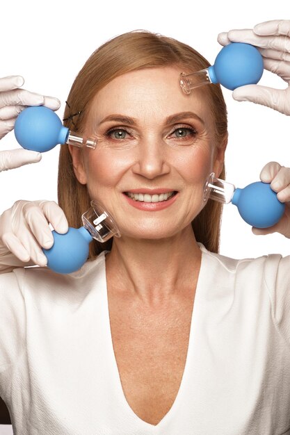 Photo portrait of a beautiful elderly woman in a white shirt with classic makeup and blond hair with massage tools in hands