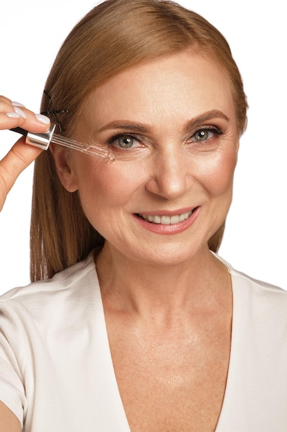 Photo portrait of a beautiful elderly woman in a white shirt with cosmetic face oil in her hands