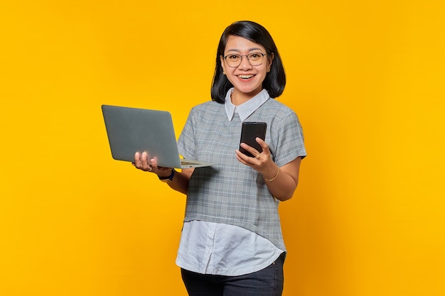 Portrait of beautiful excited Asian woman holding laptop and cell phone on blue background