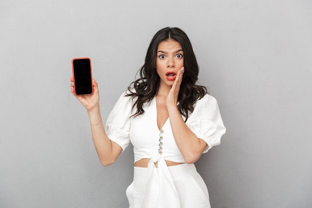 Portrait of a beautiful excited young brunette woman wearing summer outfit standing isolated over gray wall, showing blank screen mobile phone