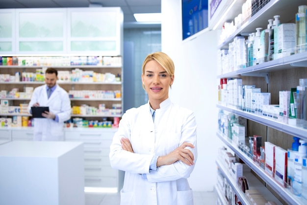 Photo portrait of beautiful female blonde pharmacist standing in pharmacy shop or drugstore with her arms crossed.