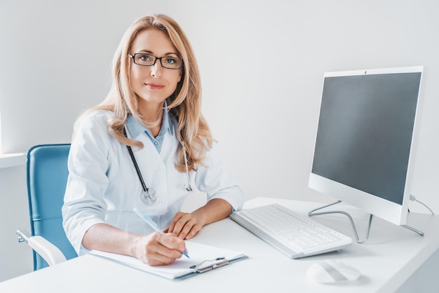 Portrait of beautiful female doctor with desktop pc and clipboard sitting at desk in clinic