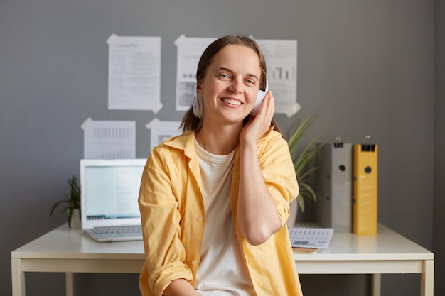 Portrait of beautiful female freelancer sitting in office while having break listening to music or podcast wearing casual clothing looking at camera holding headphones