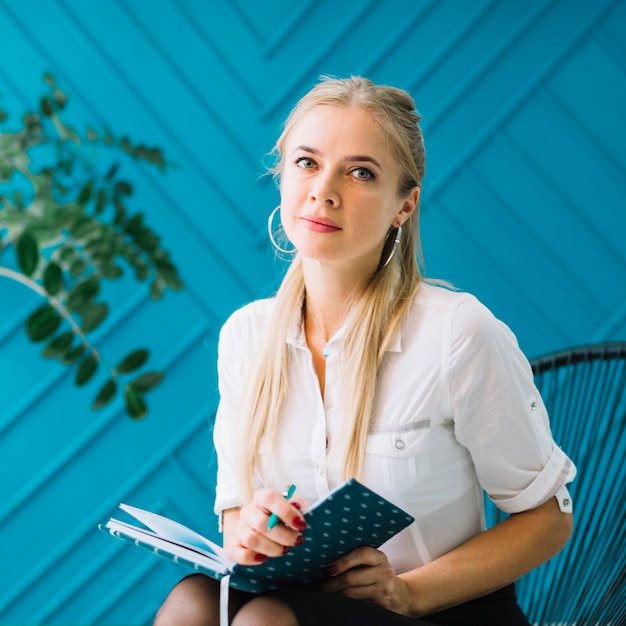 Foto ritratto di bello psicologo femminile con il diario e penna che si siedono sulla sedia davanti alla parete blu che guarda alla macchina fotografica