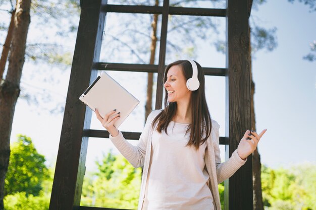 Portrait of beautiful fun woman wearing light casual clothes. Pretty girl holding tablet pc computer, listening music in headphones in city park in street outdoors on spring nature. Lifestyle concept.