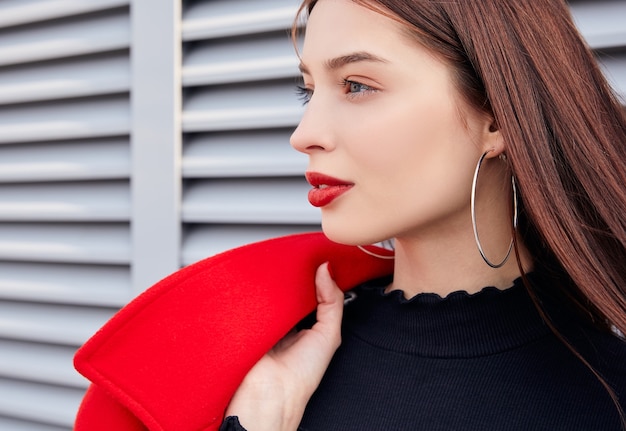 Photo portrait of beautiful girl in black top and big earings holding over her shoulder a red coat on grey street wall background