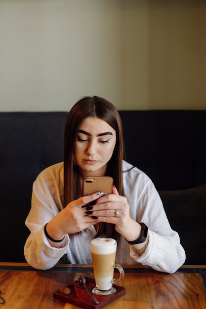 彼女の携帯電話でカフェで熱いお茶やコーヒーを飲む美しい少女の肖像画。