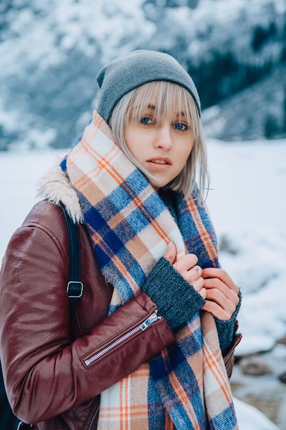 Portrait of a beautiful girl in a hat and scarf on a background of snowcappe A look at the camera