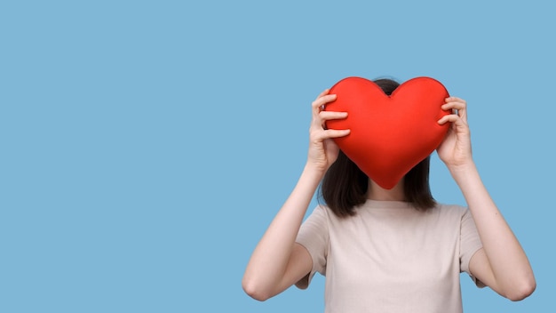 Portrait of a beautiful girl holding a red pillow in the shape of a heart instead of a face on a blue background