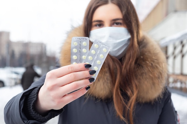 Portrait of a beautiful girl in a medical protective mask in holding yellow pills in her hands for colds and flu Winter street portrait of a woman under snowfall