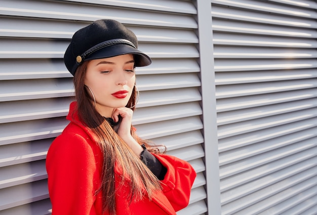 Portrait of beautiful girl in red coat and black hat posing with grey street wall background