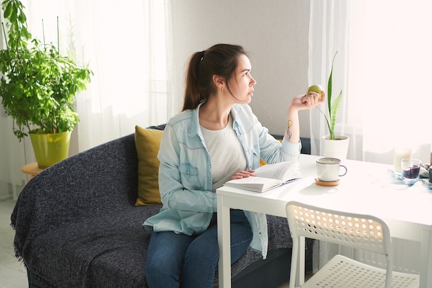 Portrait of beautiful girl sitting at the table, reading a book and eating an apple