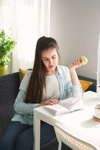 Portrait of beautiful girl sitting at the table, reading a book and eating an apple