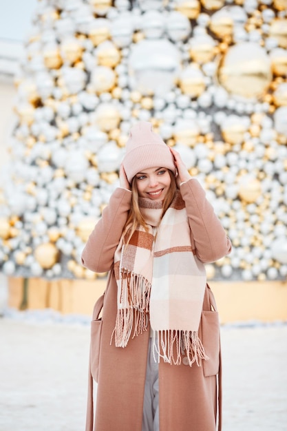 Photo portrait of beautiful girl in stylish winter beige outfit in front of christmas tree outdoors.
