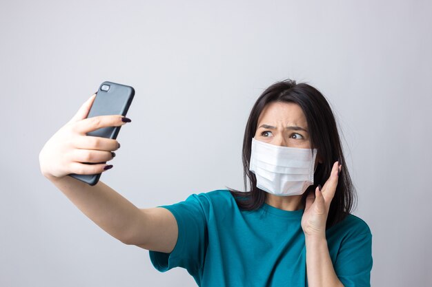 Portrait of a beautiful girl taking a selfie on a gray background wearing a medical mask.