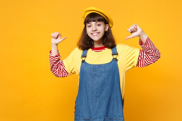 Portrait of beautiful girl teenager in french beret, denim sundress pointing thumbs on herself isolated on yellow background in studio. People sincere emotions, lifestyle concept. Mock up copy space.