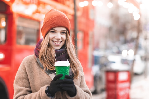 Ritratto bella ragazza in abiti caldi in piedi in strada con un bicchiere di carta di caffè sullo sfondo bokeh di fondo