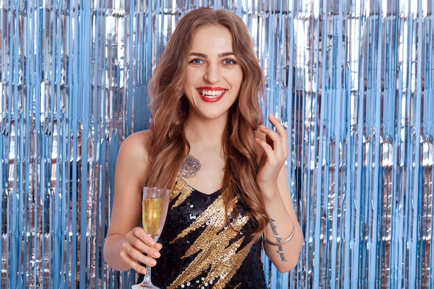 Photo portrait of beautiful girl with happy charming smile on her face, has bright make-up and hairstyle, holding glass of wine in her hands