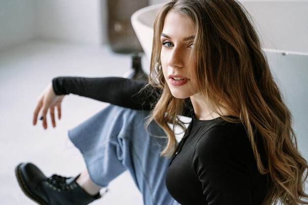 Portrait of a beautiful girl with light brown long hair in a black t-shirt, jeans and rough boots. Stylish bathroom