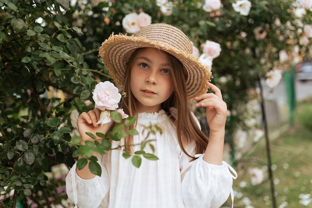 Portrait of a beautiful girl with long hair and freckles in a straw hat outside in the summer near roses