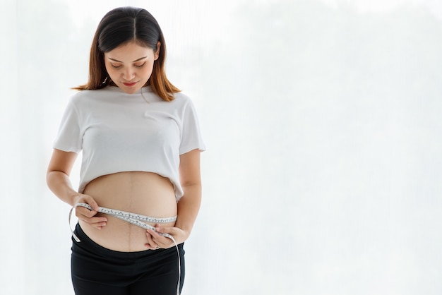 Portrait of beautiful happy Asian pregnant woman wearing a casual dress smiling standing use a measuring tape measure her bell while looking at a camera.