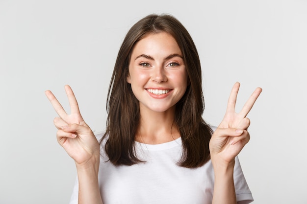 Portrait of beautiful and happy brunette girl smiling and showing peace gestures, standing white.