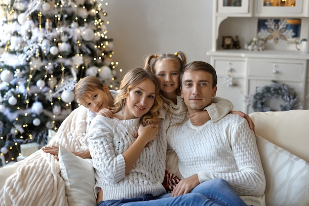 Portrait of beautiful happy family with two children in white sweaters sitting on sofa in festively decorated room with a Christmas tree