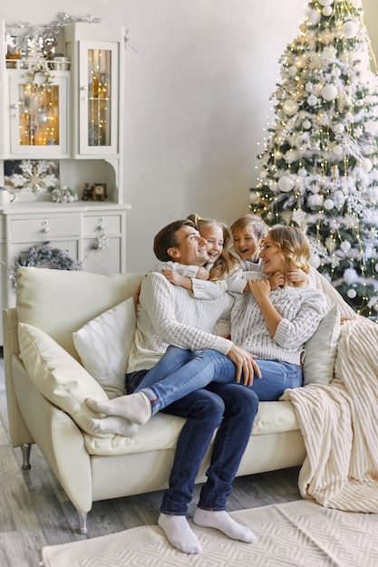 Portrait of beautiful happy family with two children in white sweaters sitting on sofa in festively decorated room with a Christmas tree