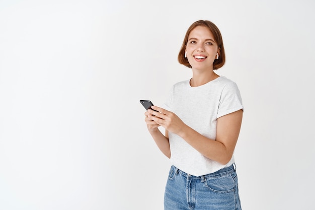 portrait of beautiful happy girl holding smartphone screen and smiling, listening music in wireless headphones, standing against white wall
