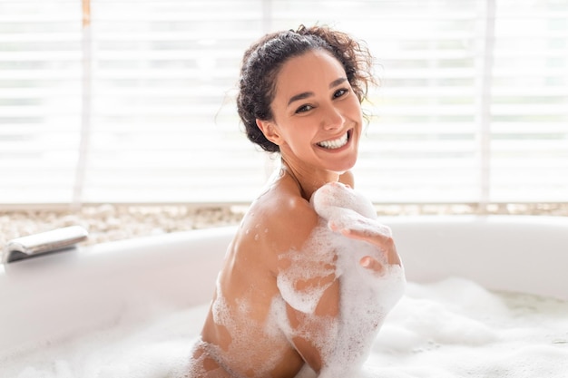 Portrait of beautiful happy millennial woman sitting in hot bubble bath covered in foam at hotel