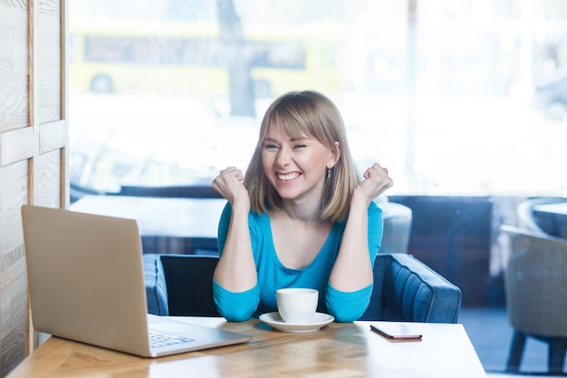 Portrait of beautiful happy satisfied positive young girl freelancer with blonde hair in blue t-shirt sitting in cafe and triumphing with raised arms and celebrating her victory, looking at camera.