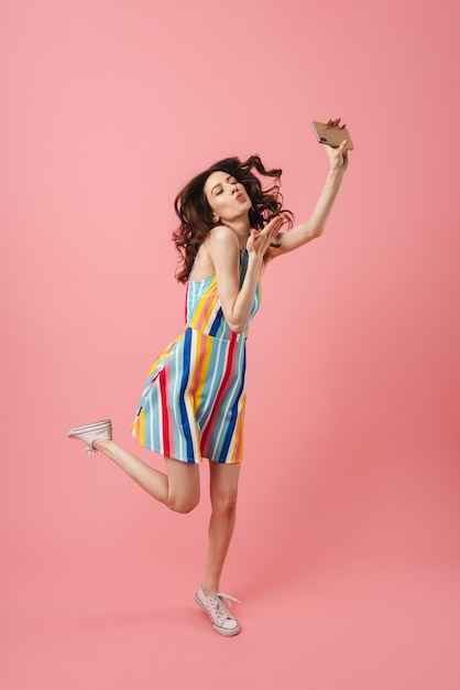 Portrait of beautiful happy smiling pleased young cute woman posing isolated over pink wall take a selfie by mobile phone blowing kisses