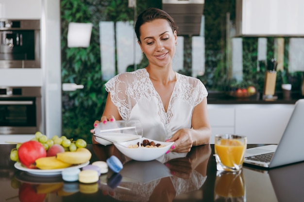 The portrait of a beautiful happy woman pouring milk into a bowl with cereal for breakfast with a laptop on the table