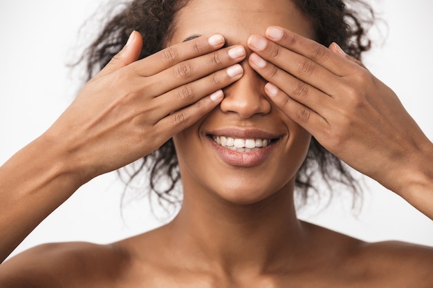 portrait of a beautiful happy young african woman posing isolated over white wall covering eyes with hands.