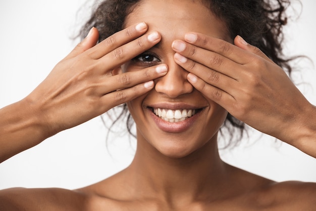 portrait of a beautiful happy young african woman posing isolated over white wall covering eyes with hands.