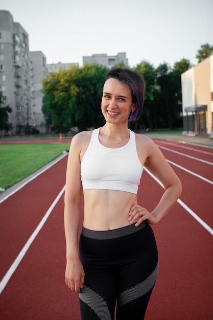 Foto ritratto bella felice giovane donna corridore atleta in maglietta bianca e abbigliamento sportivo allo stadio guardando la telecamera e sorridente nella corsa mattutina e allenamento allo stadio all'aperto