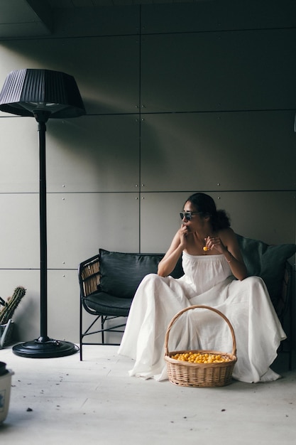 Portrait of a beautiful indian woman in white dress and sunglasses sitting on the sofa with a basket of yellow sweet cherry