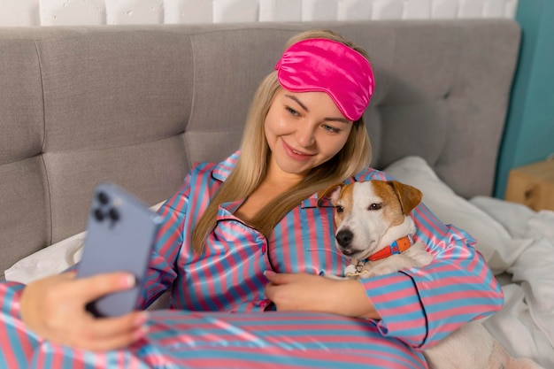 Portrait of beautiful joyful woman in pajamas hugging dog using mobile phone for selfie smiling