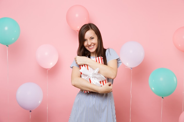 Portrait of beautiful laughing young woman in blue dress holding and hugging red box with gift present on pink background with colorful air balloons. Birthday holiday party, people sincere emotions.